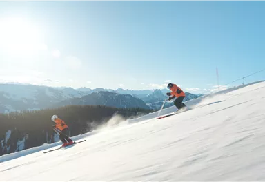 Zwei Skifahrer fahren die Piste hinunter. Im Hintergrund sind schneebedeckte Berge und ein klarer blauer Himmel zu sehen.