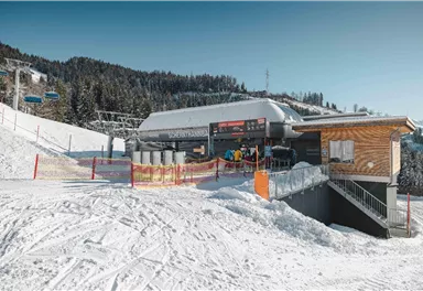 Eine moderne Skihütte in einer schneebedeckten Landschaft. Im Hintergrund sind Skilifte und Wälder sichtbar.