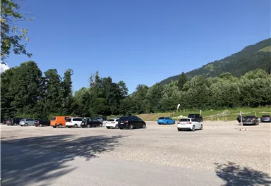 A large parking lot, partially filled, surrounded by trees and mountains. The sky is clear and blue.