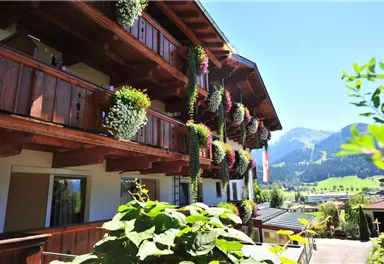 A traditional wooden building with flower baskets and a magnificent mountain view. The sky is clear and blue, the surroundings are green and inviting.