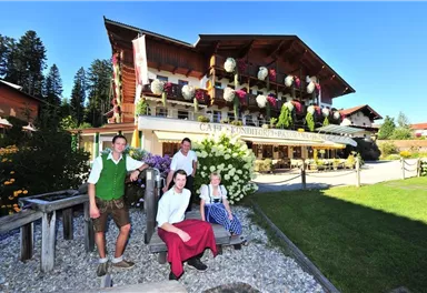 A traditional café in Bavarian style with blooming flowers and a well-kept garden. Four people in traditional costumes stand in front of the building and smile friendly.