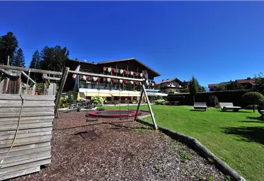 A beautiful garden with a swing and lots of green grass. In the background, there is a building with balconies and sun umbrellas.