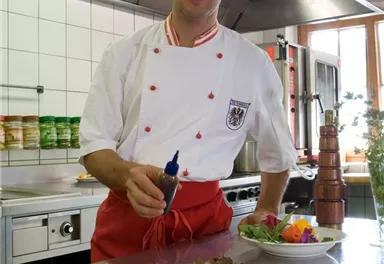 A chef stands in a modern kitchen and presents an artistically arranged plate with salad and vegetables. In the background, kitchen appliances and other dishes are visible.