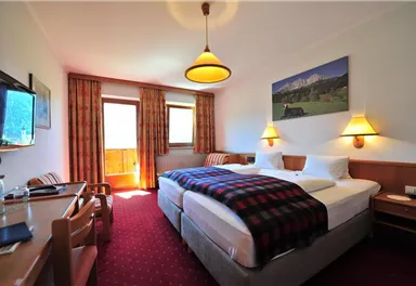 A cozy hotel room with two beds and a table. The windows let in plenty of daylight and there is a view of the mountains.