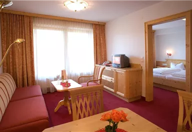 A cozy living area with a sofa, a table, and a television. In the background, a bedroom with a double bed is visible.