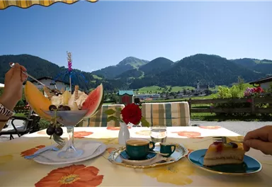 A set table with a large bowl of fruit salad and a piece of cake. In the background, green mountains and a blue sky can be seen.