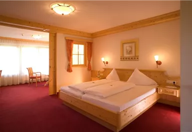 A cozy hotel room with a large bed and wooden details. There is a sunny seating area by a window and a cozy atmosphere.