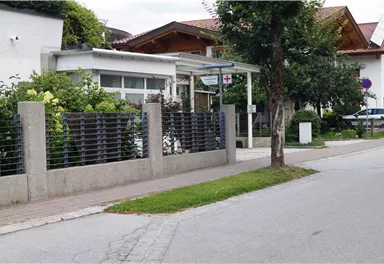 Ein modernes Gebäude an einer ruhigen Straße, umgeben von Büschen und Bäumen. Die Umgebung zeigt ein gepflegtes Wohngebiet mit Bürgersteig und Parkplätzen.