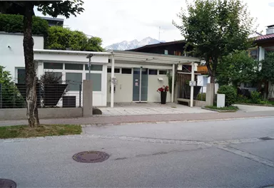 Ein modernes Gebäude mit großen Fenstern und einem Eingangsbereich. Im Vordergrund sind Bäume und eine ruhige Straße sichtbar.