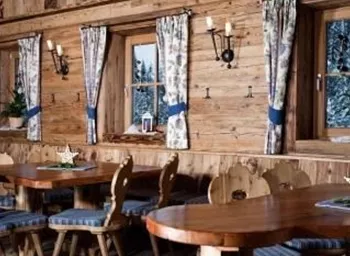 A cozy interior with wooden walls and large windows. The tables are made of wood and equipped with blue seat cushions.