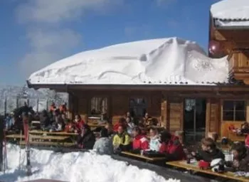A ski hut with many guests outside, surrounded by snow. The sky is blue and clear.