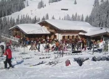 A lively ski hut area in the snow with many skiers and ski equipment. In the background, there are snow-covered trees and mountains.