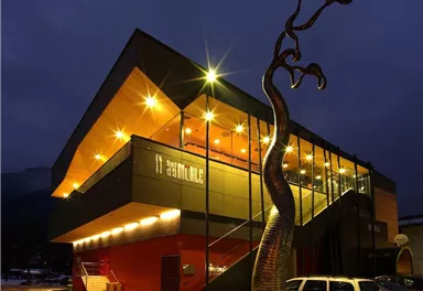 A modern building with spectacular design at night. In front of the facade stands a striking sculpture.