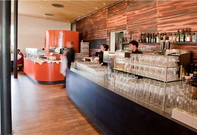 A modern restaurant interior with a long bar counter and many glasses. In the background, you can see staff serving guests.
