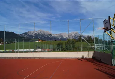 A basketball court with a red surface in the mountains. In the background, green meadows and snow-covered peaks can be seen.