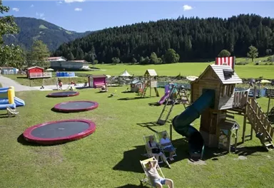 A spacious playground with trampolines, slides, and play equipment. Surrounded by green grass and mountains.