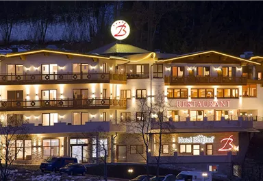 A modern hotel with warm lighting and an inviting restaurant. The facade features cozy balconies and is surrounded by trees.