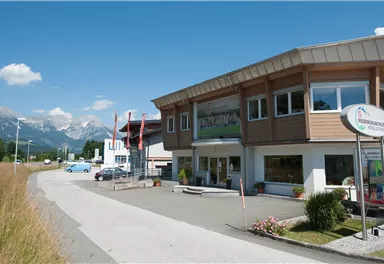 Ein modernes Gebäude mit großen Fenstern steht an einer Straße. Im Hintergrund sind Berge und ein klarer blauer Himmel sichtbar.