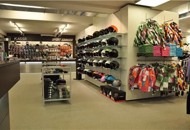 Ein Sportgeschäft mit einer Vielzahl von Sportartikeln. An den Wänden hängen Bekleidung und Helme, während die Kasse im Hintergrund sichtbar ist.