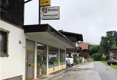 Ein Elektrogeschäft mit dem Namen "Salvenmoser" an einer ruhigen Straße. Im Hintergrund sind typische alpine Gebäude und ein bewölkter Himmel zu sehen.