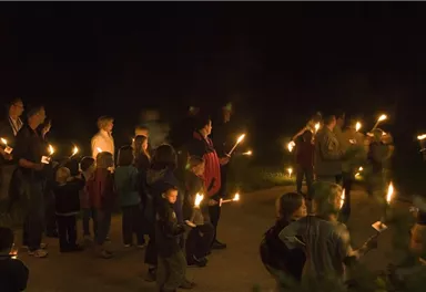 Eine Gruppe von Menschen mit Fackeln versammelt sich nachts. Sie stehen auf einem Weg und scheinen an einer Feier oder Zeremonie teilzunehmen.