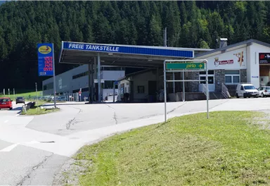 Eine Tankstelle mit dem Schild "Freie Tankstelle" in einer grünen Berglandschaft. Im Vordergrund sind Autos und Wegweiser zu sehen.