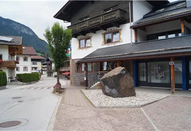 A quiet street with traditional buildings and a large stone in the middle. In the background, green mountains are visible.