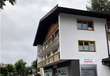 Ein zweigeschossiges Gebäude mit einem Friseursalon und einem klaren Straßenverlauf. Über dem Eingang stehen Blumen und der Himmel ist bewölkt.
