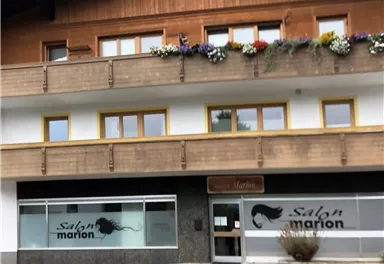 Ein Friseursalon namens "Salon Marion" mit großen Fenstern und einem Holzgebäude. Es gibt Blumenkästen an den Fenstern und eine angrenzende Straße.