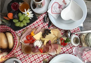 Ein gedeckter Tisch mit einer Auswahl an Wurst, Käse, Gemüse, und Früchten. Dazu gibt es Brot, Getränke und dekorative Elemente auf einem karierten Tuch.