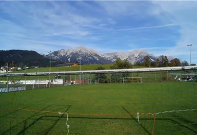 A soccer field with green grass and a mountain landscape in the background. The mountains are partly covered with snow and the sky is clear.