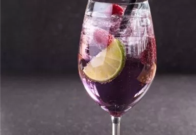 An elegant glass with purple drink, garnished with lime slices and berries. The drink has a colorful, sparkling character and a straw.