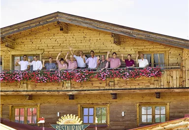 Eine Gruppe von Menschen steht auf dem Balkon eines Holzhauses und winkt fröhlich. Der Balkon ist mit bunten Blumen geschmückt.
