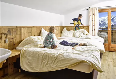 Ein gemütlicher Schlafraum mit zwei großen Betten und einer Holzwand. Drei Kinder spielen auf den Betten und genießen die entspannte Atmosphäre.