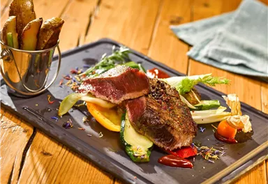 Ein saftiges Stück Fleisch mit buntem Gemüse und einer Beilage von Ofenkartoffeln. Das Gericht wird auf einem schwarzen Teller präsentiert und sieht sehr appetitlich aus.