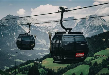 A cable car glides over green meadows and majestic mountains. In the background, tall, snow-capped peaks are visible.