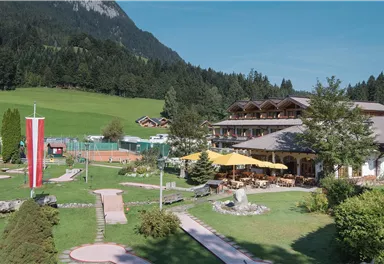 A picturesque hotel in the mountains with a beautiful garden and a mini-golf course. The clear sky and the green landscape create a relaxed atmosphere.