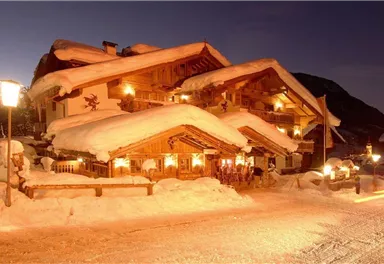 Ein traditionelles Chalet im Schnee mit schneebedecktem Dach. Die warme Beleuchtung schafft eine gemütliche Atmosphäre in der winterlichen Landschaft.