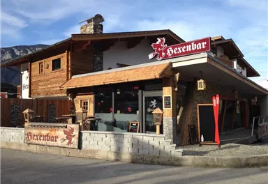 Eine rustikale Bar namens "Hexenbar" mit einem einladenden Außenbereich. Die Fassade besteht aus Holz und Stein, umgeben von Bergen.