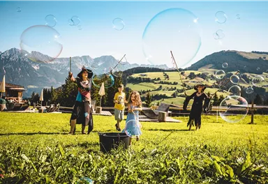 Eine fröhliche Szene im Freien mit Kindern, die Seifenblasen fangen. Im Hintergrund sind Berge und eine grüne Wiese zu sehen.