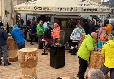 Ein belebter Après-Ski-Bereich mit vielen Menschen, die um Holzstämme und einen Grill versammelt sind. Im Hintergrund ist ein Chalet mit einem großen Sonnenschirm zu sehen.