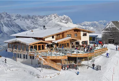 Eine gemütliche Berghütte im Schnee, umgeben von beeindruckenden Bergen. Viele Menschen genießen die Wintersonne auf der Terrasse.