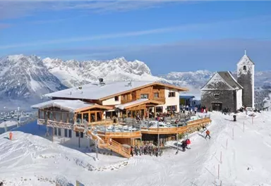 Ein schneebedecktes Bergdorf mit einem modernen Restaurant und einer alten Kirche. Die Umgebung ist von majestätischen Bergen und klarem Himmel umgeben.