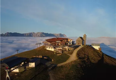 Eine Berghütte liegt hoch über den Wolken mit Blick auf die Berge im Hintergrund. Die Szenerie ist friedlich und bietet eine atemberaubende Aussicht.