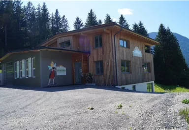 Ein modernes Holzhaus in einer bergigen Umgebung. Der Eingang ist freundlich gestaltet und von Bäumen umgeben.