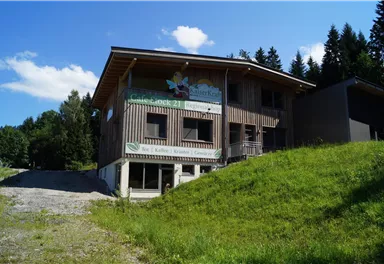 Ein modernes Holzgebäude in einer naturnahen Umgebung. Im Hintergrund sind Bäume und ein blauer Himmel zu sehen.