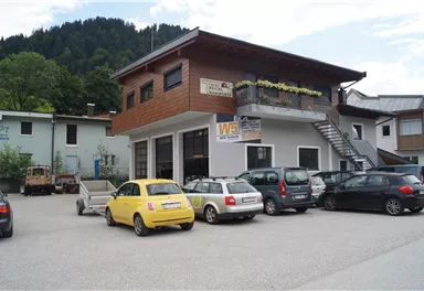 Ein modernes Gebäude mit Holzverkleidung in einer ländlichen Umgebung. Auf dem Parkplatz sind mehrere Autos geparkt, darunter ein gelbes Auto.