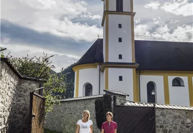 Zwei Personen gehen auf einem Weg vor einer Kirche. Der Himmel ist klar und die Sonne scheint.