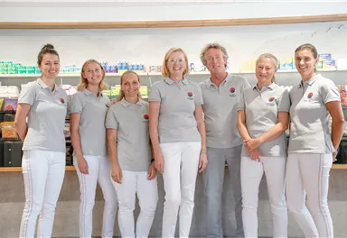 Ein Team von sieben Personen steht vor einem Regal mit Gesundheitsprodukten. Alle tragen graue Poloshirts und weiße Hosen.
