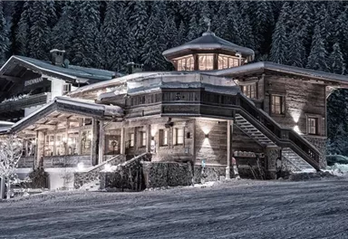 Ein gemütliches Holzhaus im Schnee, umgeben von hohen Tannenbäumen. Die Umgebung ist winterlich und friedlich.